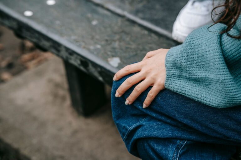 Mains posées sur un genou, pull en laine vert d'eau, personne assise sur un banc